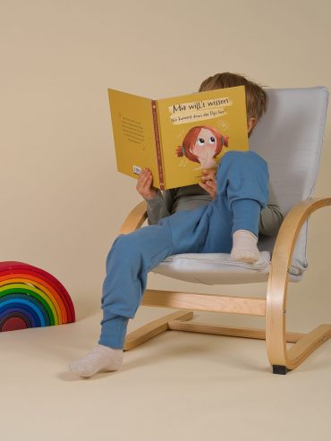 Kind sitzt im Kindersessel mit Windelweg Hose und ließt das Buch "Mia will's wissen" von Aylin Knapp