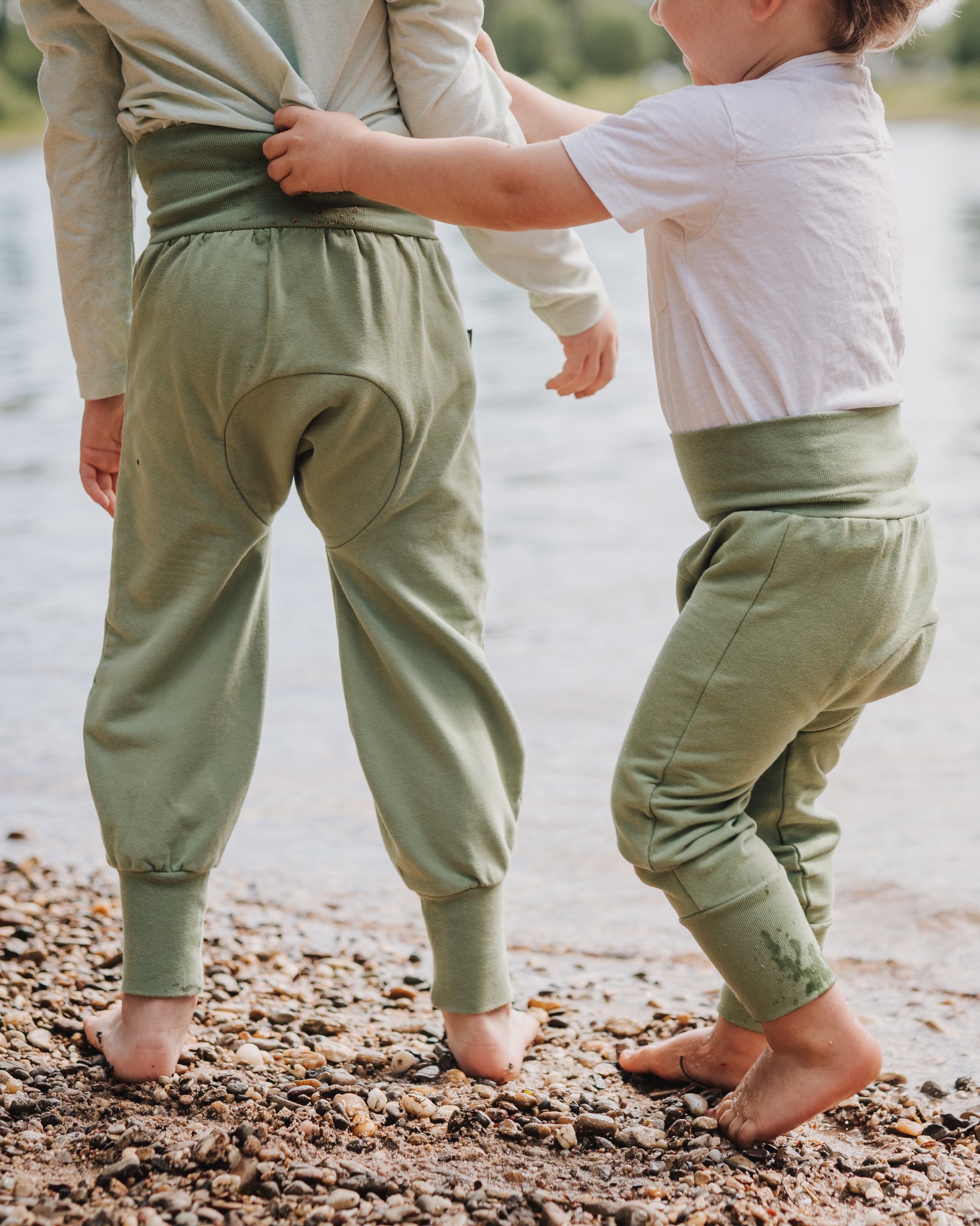 Zwei Kinder spielen am Flussrand und beide tragen die Windelweg Hose Auengrün, die sie beim Trockenwerden begleitet